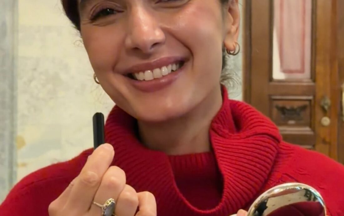 Smiling person in a red sweater holding lipstick and a compact mirror, as if applying makeup.