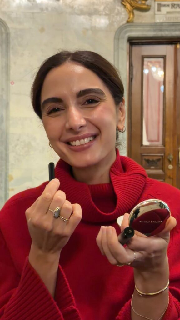 Smiling person in a red sweater holding lipstick and a compact mirror, as if applying makeup.