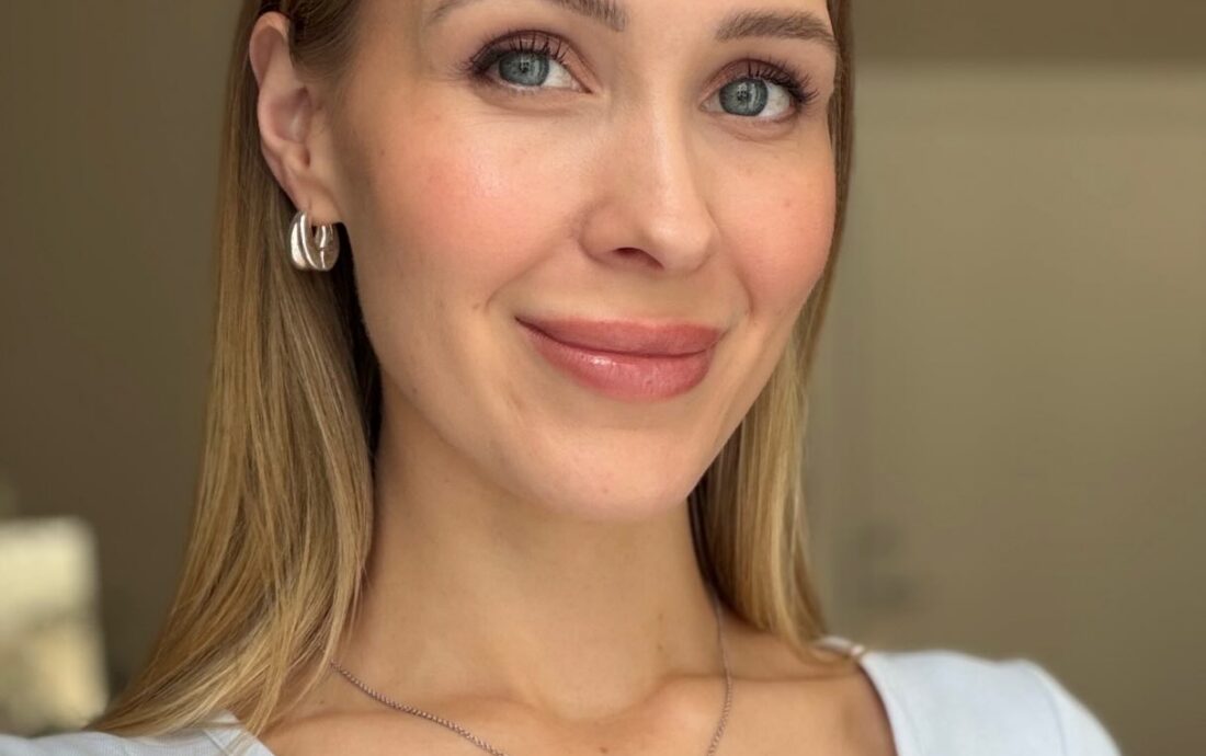 Close-up portrait of a smiling woman with blonde hair and blue eyes, wearing a light blue top and silver jewelry.