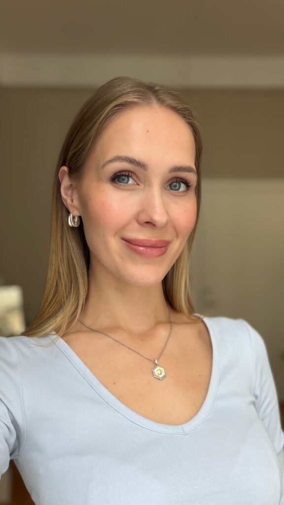 Close-up portrait of a smiling woman with blonde hair and blue eyes, wearing a light blue top and silver jewelry.