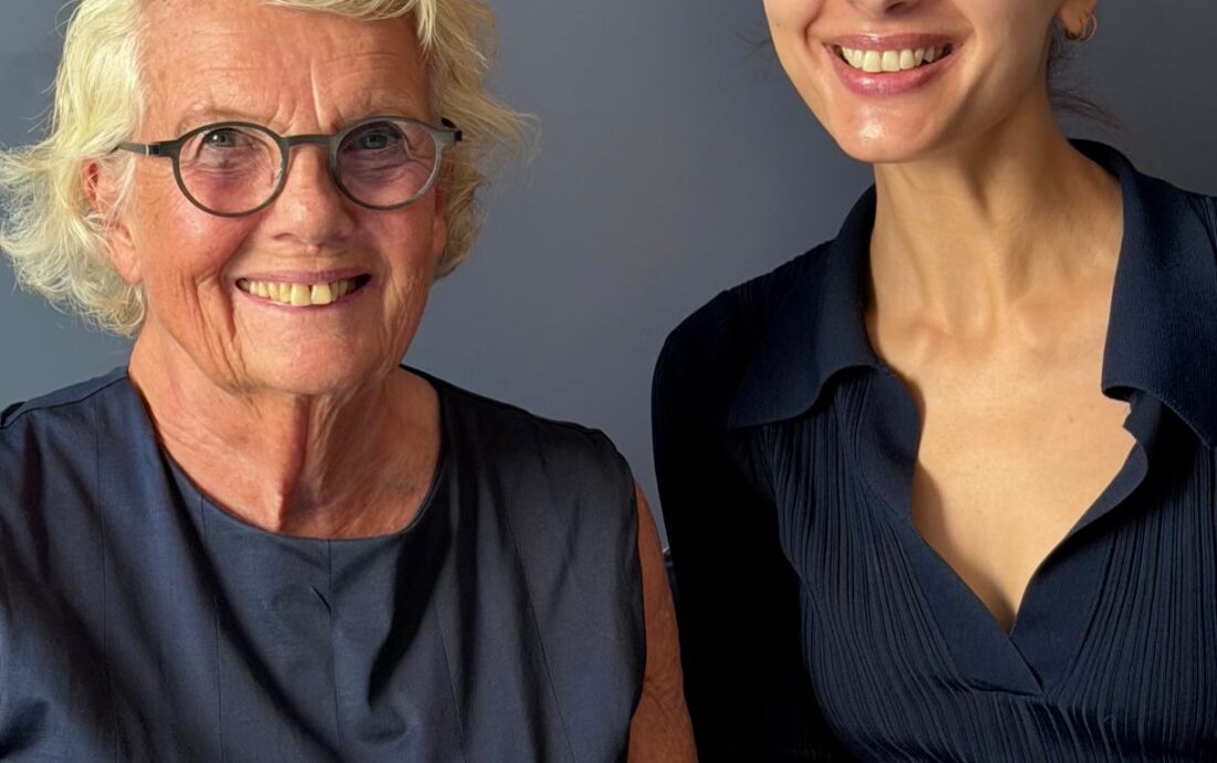 Two women smile at the camera: a senior with glasses on the left and a younger woman on the right, both in navy clothing, posing with Noble Panacea skincare products on a table.