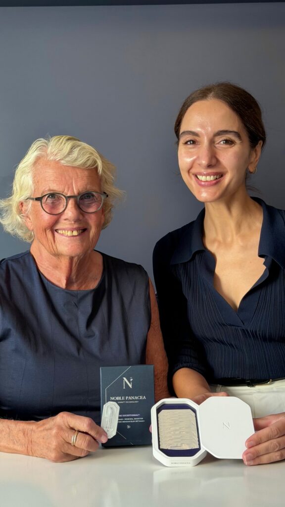Two women smile at the camera: a senior with glasses on the left and a younger woman on the right, both in navy clothing, posing with Noble Panacea skincare products on a table.
