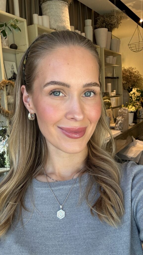 Close-up portrait of a smiling woman with blue eyes and light brown hair, wearing a silver pendant necklace; shop shelves and decor behind her.