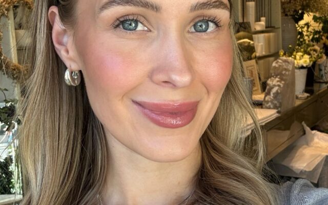 blogg-red-3992363185846362686-frame0 - Gimle Parfymeris blogg Close-up portrait of a smiling woman with blue eyes and light brown hair, wearing a silver pendant necklace; shop shelves and decor behind her.