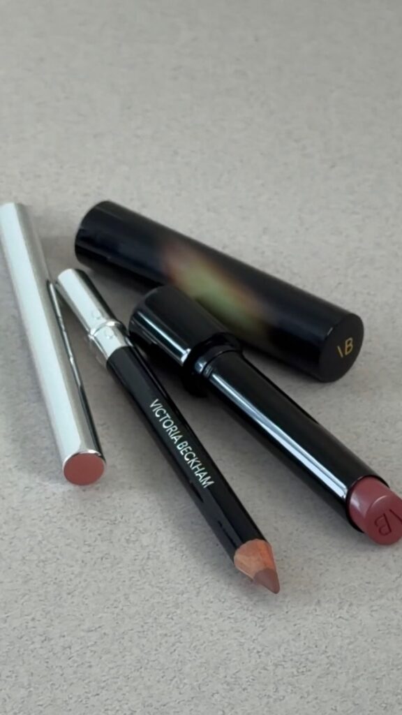 Close-up of makeup items on a light surface: a Victoria Beckham lip pencil, a black lip liner, a black lipstick with a pinkish-red shade, and a silver-toned lip product with a cap nearby.