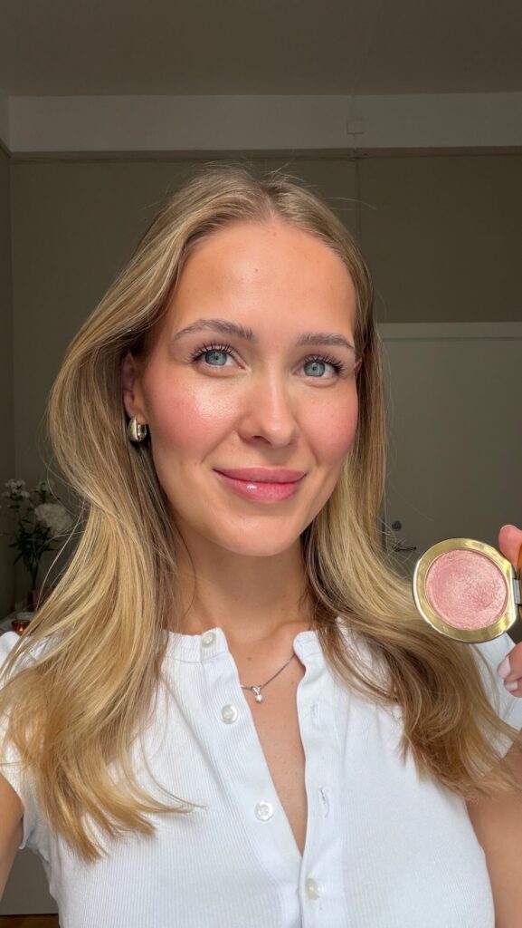 Portrait of a smiling woman with blue eyes and long blonde hair, holding a pink makeup compact toward the camera.
