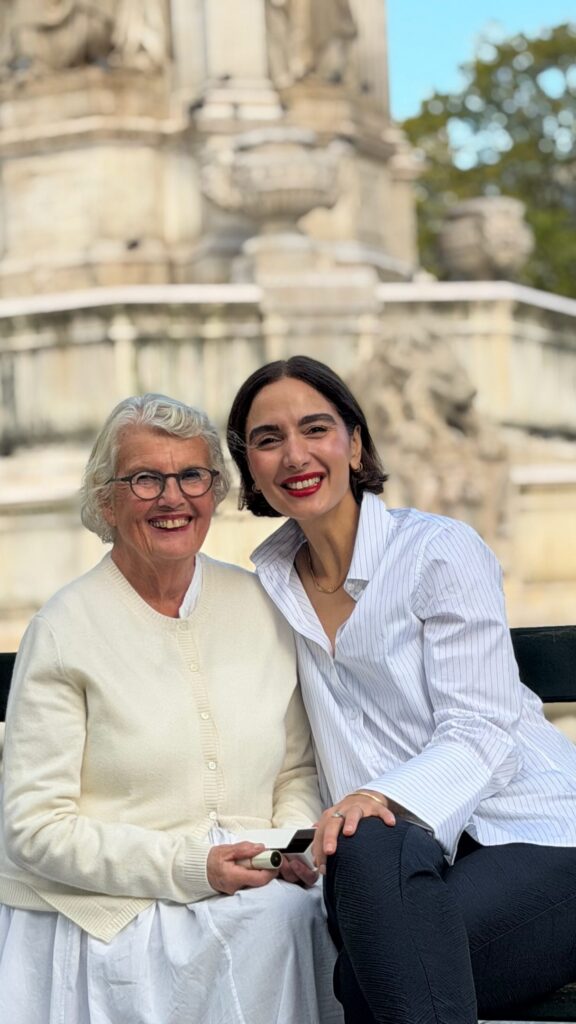 Two smiling women posing together on a bench outdoors, with a historic stone structure in the background.