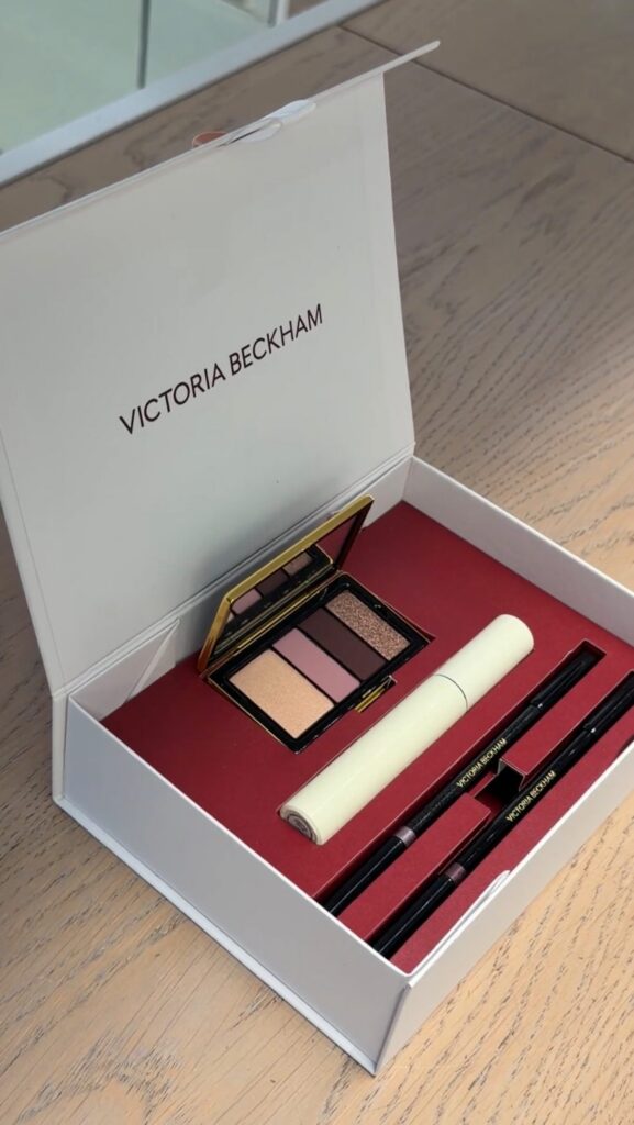 Opened Victoria Beckham makeup set in a white box, showing a peach-pink eyeshadow palette and black pencils on a red interior surface.